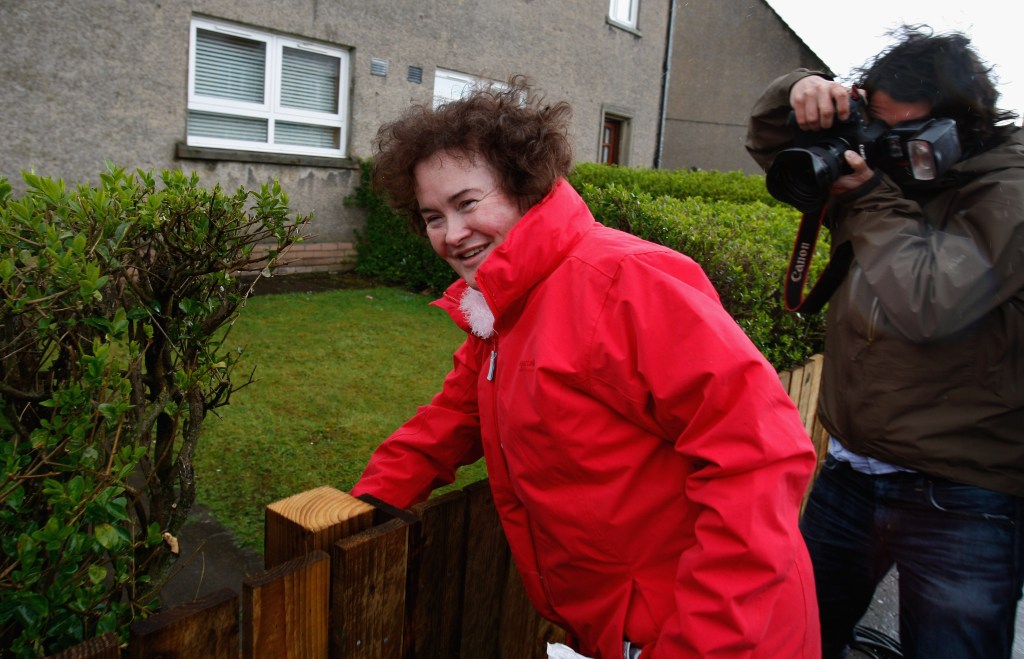Susan Boyle leaning against a wooden fence, smiling at the camera.
